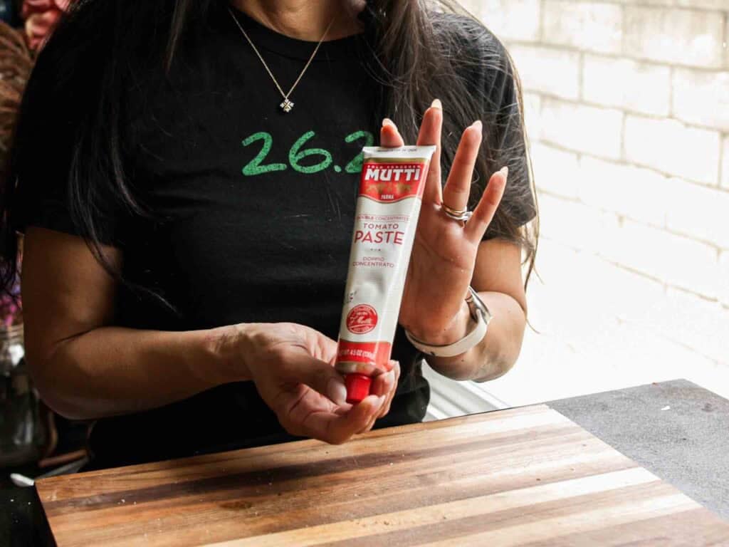 A person wearing a black shirt that says “26.2” holds a tube of Mutti tomato paste over a wooden cutting board, with a bright background visible in the distance.